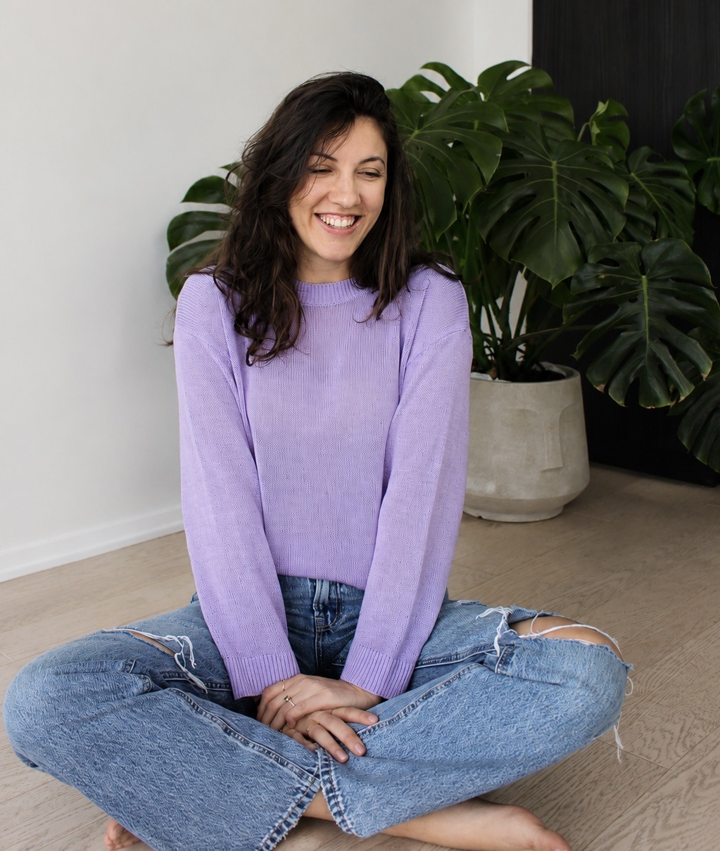 Francesca smiling in a lavender sweater by a Monstera plant